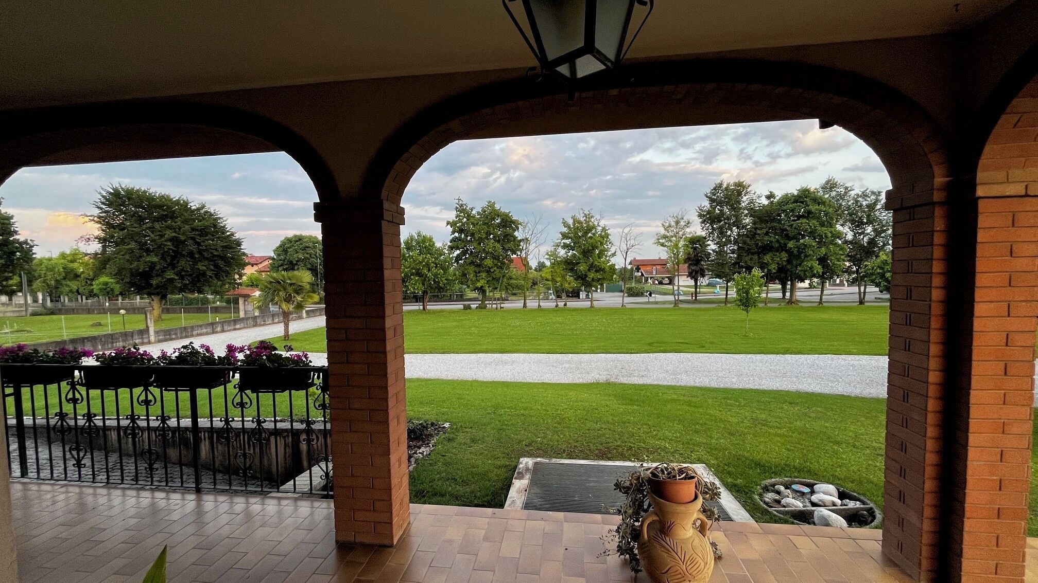 Un patio techado con columnas de ladrillo, vista a un césped verde, un sendero y un pequeño jardín con una maceta decorativa.