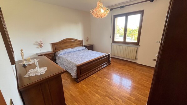 A bedroom with a wooden bed, a nightstand, a dresser, a chandelier, a window with a view, and a radiator. - Ekeko House. House surrounded by nature in the heart of Friuli's Magredi (Vivaro)