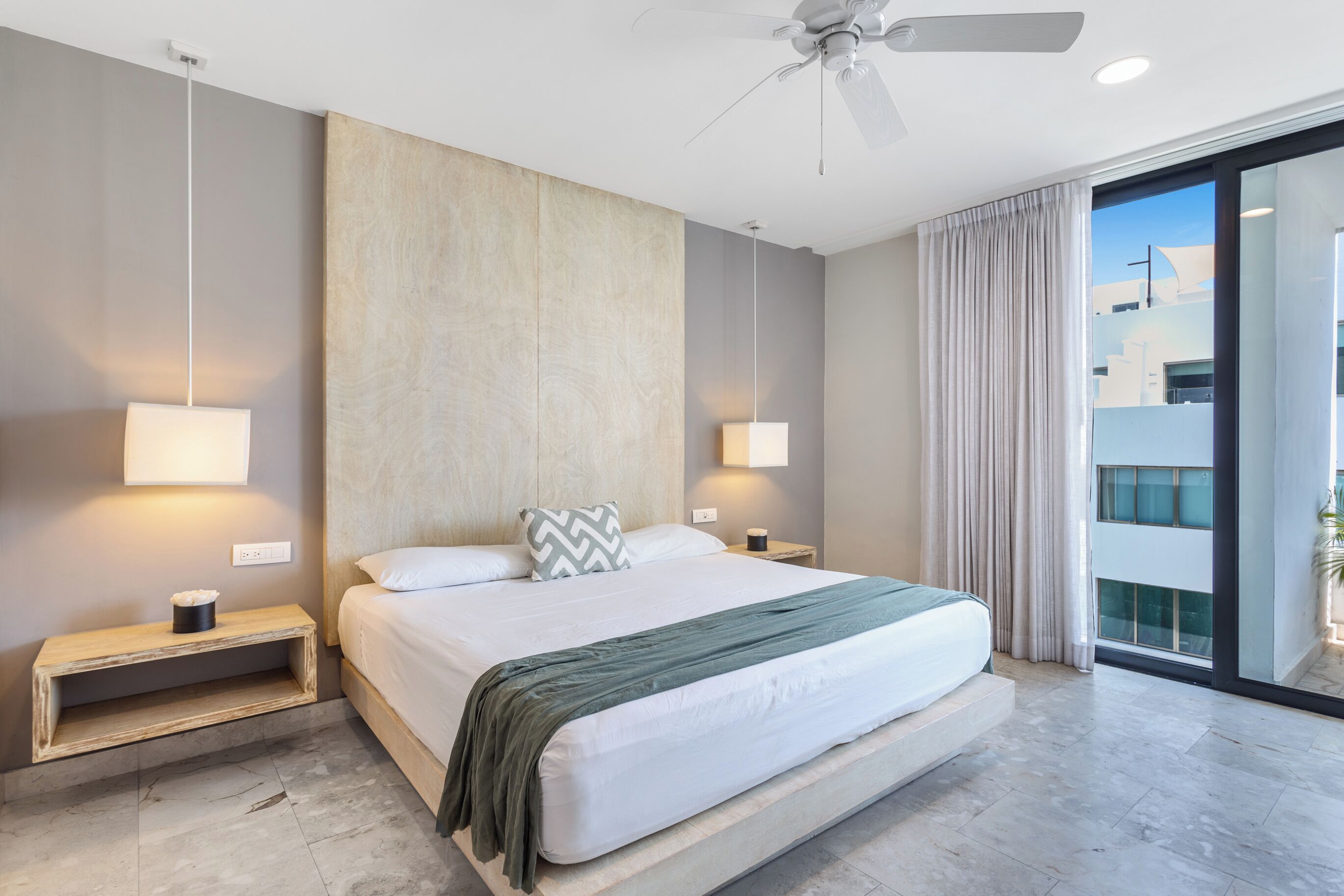 A modern hotel room with a large bed, bedside lamps, a ceiling fan, and a view through a glass door.