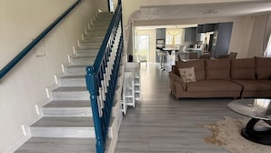 A spacious living area with a staircase, a sofa, and a kitchen in the background. - Bajan Bliss Townhouse ✈️ | 5–10 Min to Airport & Shopping. Modern and Spacious (St Philip)