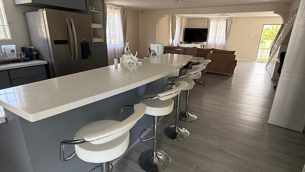 A modern kitchen with a central island, bar stools, stainless steel appliances, and a large window. - Bajan Bliss Townhouse ✈️ | 5–10 Min to Airport & Shopping. Modern and Spacious (St Philip)