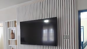 A flat-screen TV mounted on a wall with vertical slats, a red entertainment unit below, and a white shelving unit to the left. - Bajan Bliss Townhouse ✈️ | 5–10 Min to Airport & Shopping. Modern and Spacious (St Philip)