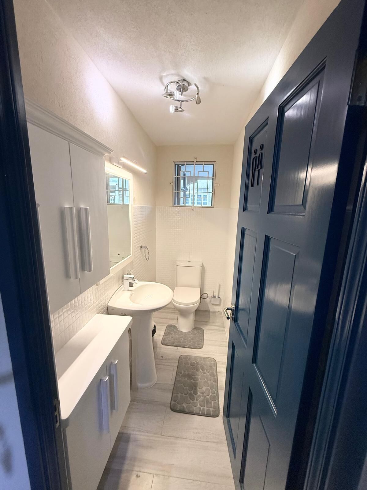 A narrow bathroom with a toilet, sink, and mirror. There is a window above the toilet and a door on the right.
