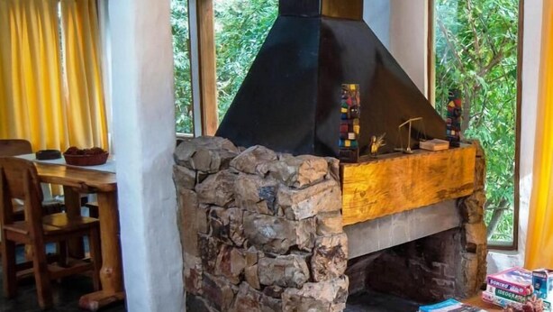 A stone fireplace with a black chimney, a dining table with chairs, and a window with a view of greenery. - Lelikelen Cabañas del Bosque (Villa La Angostura)