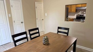 Comedor con mesa de madera, cuatro sillas, un florero y una cocina visible a través de una puerta abierta.