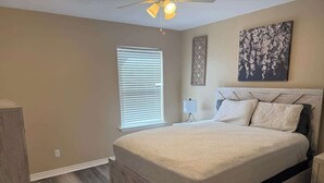 Un dormitorio con cama, mesita de noche, un cuadro en la pared, ventilador de techo y ventana con persianas.