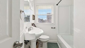 Ein Badezimmer mit weißem Waschbecken, Toilette und Badewanne, einem Fenster mit Jalousien sowie einer Tür mit silbernem Griff.