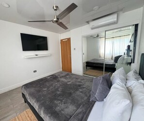 A bedroom with a bed, a TV mounted on the wall, a ceiling fan, and a mirror. - Oceanview Stay | Beach Access | Ocho Rios (Ocho Rios)