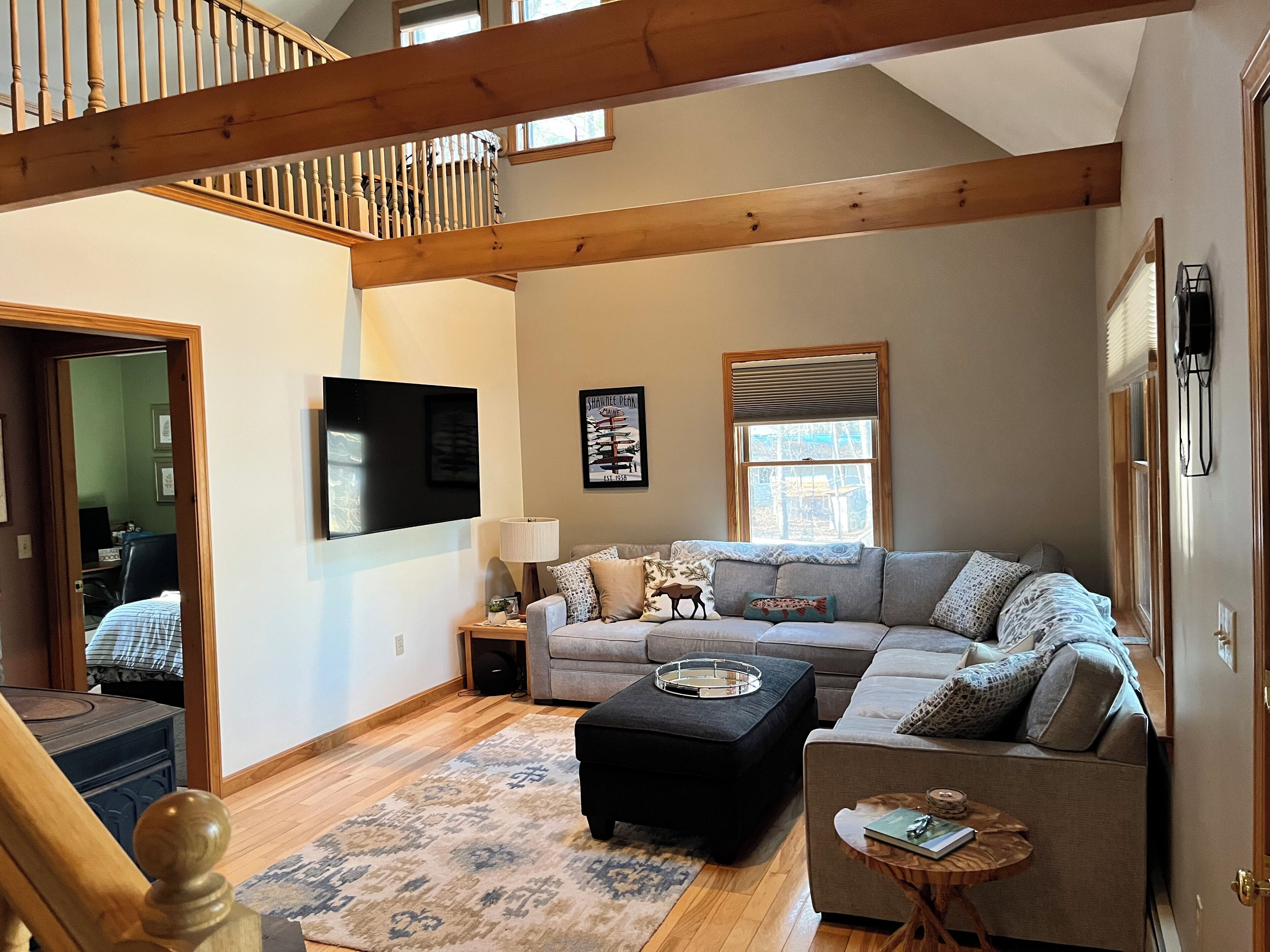 Un salon spacieux avec un grand canapé d’angle, un téléviseur à écran plat fixé au mur, un escalier en bois et une fenêtre équipée de stores.