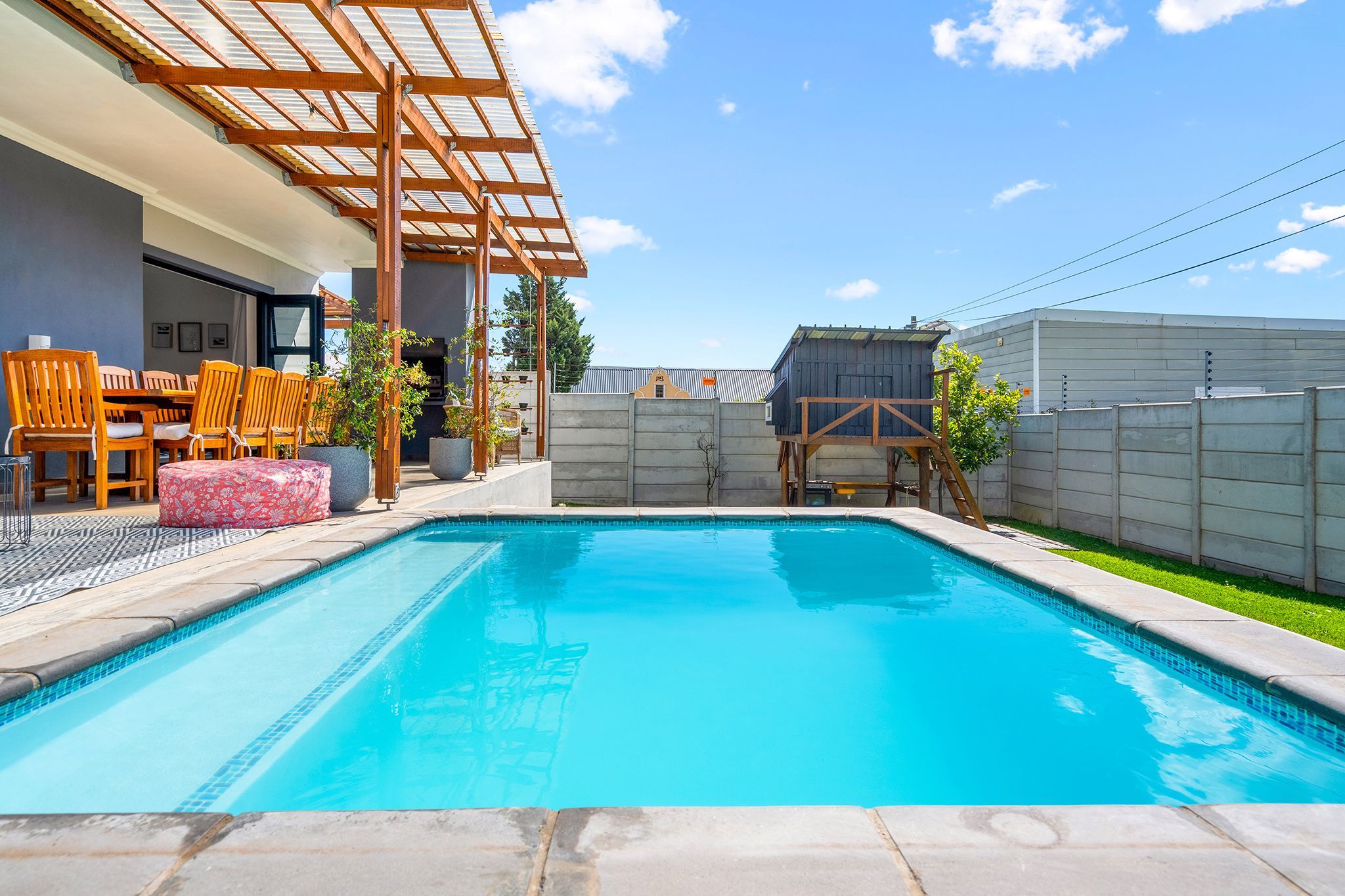 A swimming pool with a wooden deck and chairs, a covered seating area, and a fenced backyard.