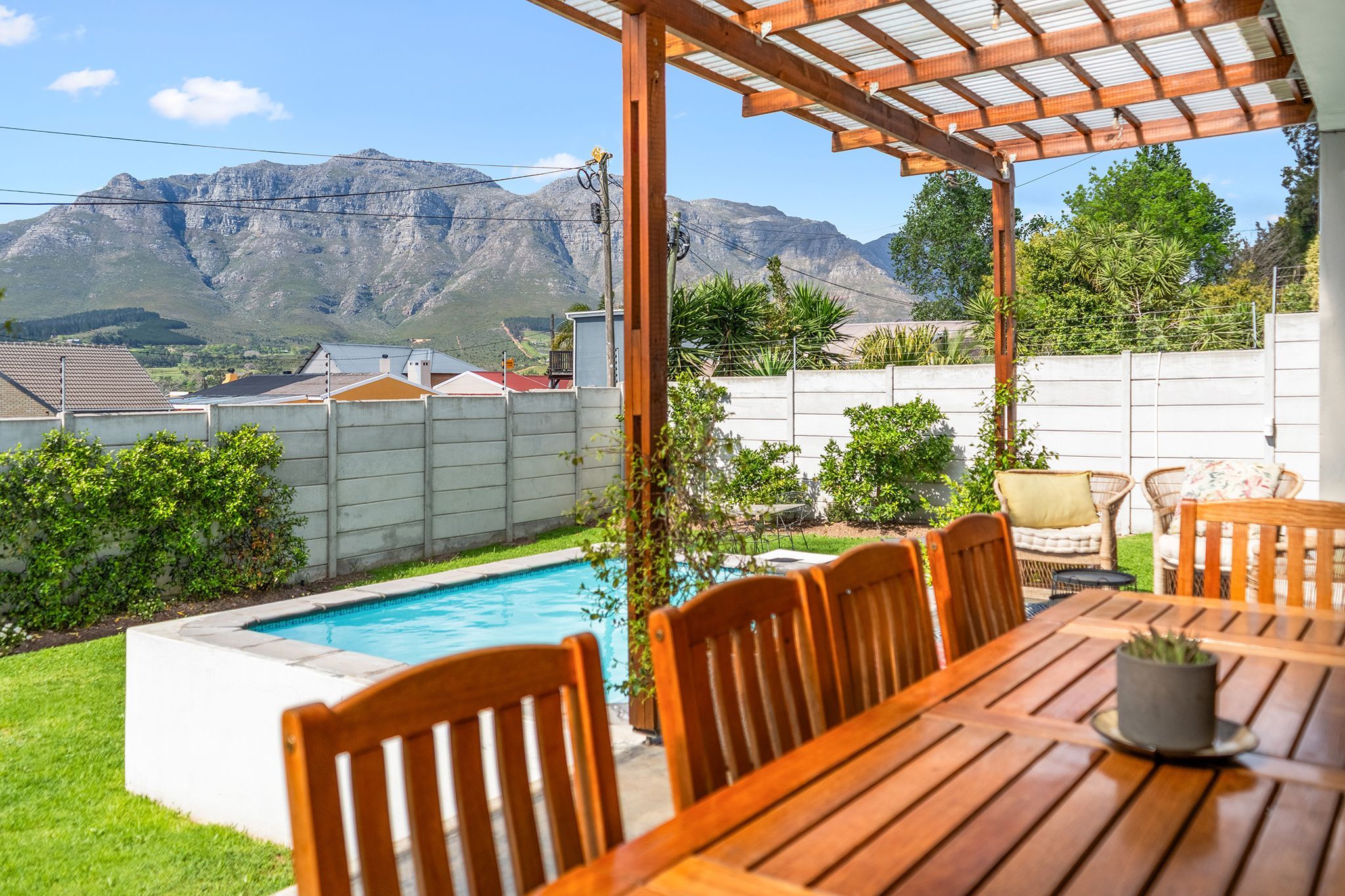 A poolside area with a wooden table and chairs, a pergola, and a view of mountains in the background.