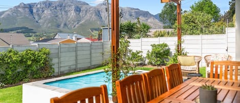 A poolside area with a wooden table and chairs, a pergola, and a view of mountains in the background.