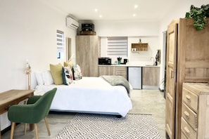 Un dormitorio moderno con cama, escritorio, una silla verde, alfombra estampada y una kitchenette al fondo.