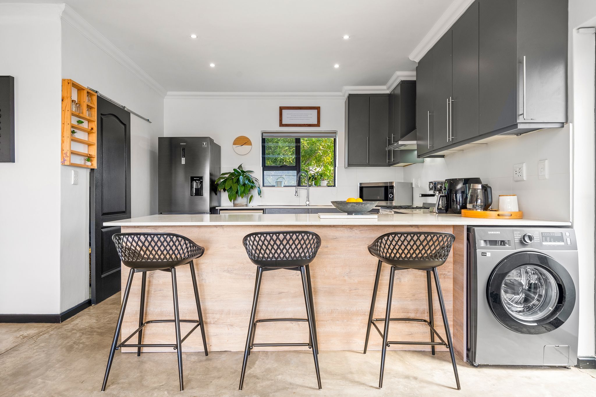 A modern kitchen with a stainless steel refrigerator, a built-in oven, a microwave, a coffee maker, and a washing machine. There are three bar stools with a lattice design.