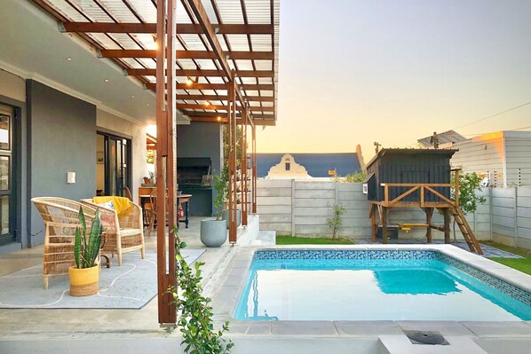 Un área de piscina moderna al aire libre con una pérgola, muebles de mimbre y una casita de juegos.