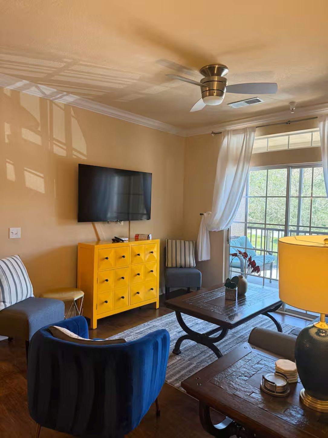 A living room with a sofa, a yellow cabinet, a blue chair, a coffee table, a TV mounted on the wall, and a window with curtains.