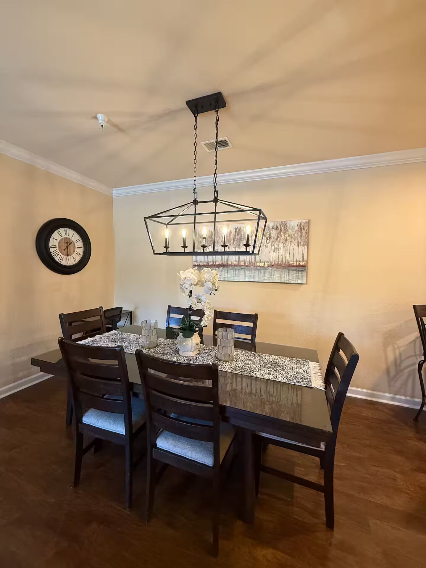 Una sala da pranzo con un tavolo in legno, sedie, un lampadario, una sveglia e un quadro appeso al muro.