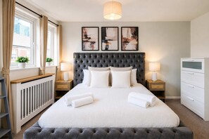 A bedroom with a large bed, two bedside tables with lamps, a radiator, a grey upholstered bench, and abstract art on the wall. - 3 Bedroom |City Centre | Modern  with Balcony (Manchester)