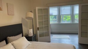 A bedroom with a bed, a bedside lamp, a window with blinds, and a door leading to an outdoor area. - Modern 2BR • 2 King Beds • Near Bakery Sq, Downtown & Universities (Pittsburgh)