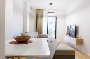 A modern living room with a white sofa, a dining table with a bowl, a TV on a wooden shelf, and a large window with curtains. - Barrikada-E – Central Apartments with French Balcony in Tirana (Tiranë)