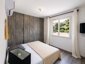 Una camera da letto con un letto, un comodino, una TV, una finestra con tende e vista sul verde circostante.