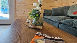 A wooden interior with a sofa, a table with a rug, a wall-mounted painting, and a window with a view.
