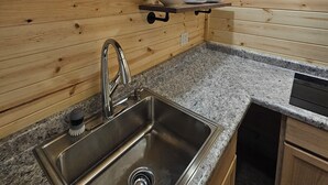 A kitchen with wooden walls, a granite countertop, a stainless steel sink, and built-in shelves with dishes.