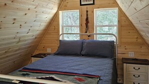A bedroom with a sloped ceiling, a bed with a white metal frame, a bedside table, and a window with a view of trees.