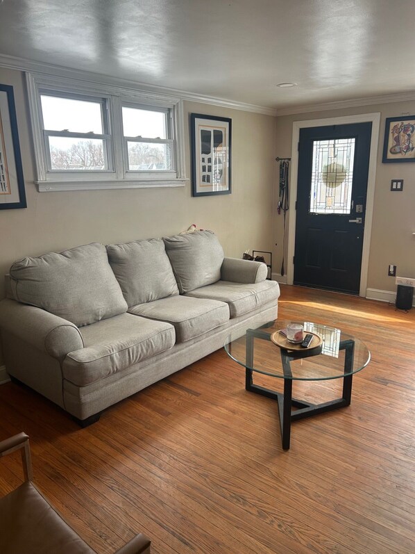 A living room with a grey sofa, a glass coffee table, wooden flooring, a black door, and a window with a view. - Serene 3-bedroom townhome in charming Drexel Hill. Ready for FIFA, MLB, PGA (Drexel Hill)