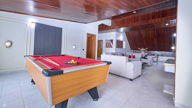 A pool table in a room with a modern design, including a wooden ceiling and a seating area with a sofa and armchairs. - THE CARRINGTON SUITES (Lagos)