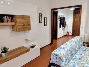 Una stanza con un divano, uno scaffale di legno con libri, una piccola pianta e una porta che dà su un'altra stanza.
