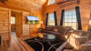 A wooden cabin interior with a sofa, a TV, and a window. - Brand New Cabin w Hot Tub, Fishing Pond, Firepit (McHenry)