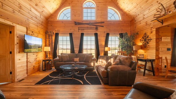 A log cabin interior with a wooden ceiling, a television, a sofa, a coffee table, and a dining area. - Brand New Cabin w Hot Tub, Fishing Pond, Firepit (McHenry)