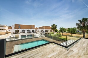 A modern outdoor swimming pool area with a glass balustrade, a wooden deck, and a view of residential buildings in the background.