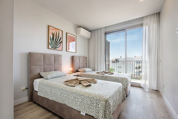 A modern hotel room with a large window, two beds, a ceiling fan, and artwork on the wall.