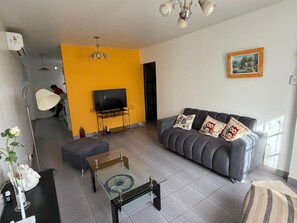 Un soggiorno con un divano, un tavolino in vetro, una TV su un mobile, una ventola a soffitto e un quadro appeso al muro.