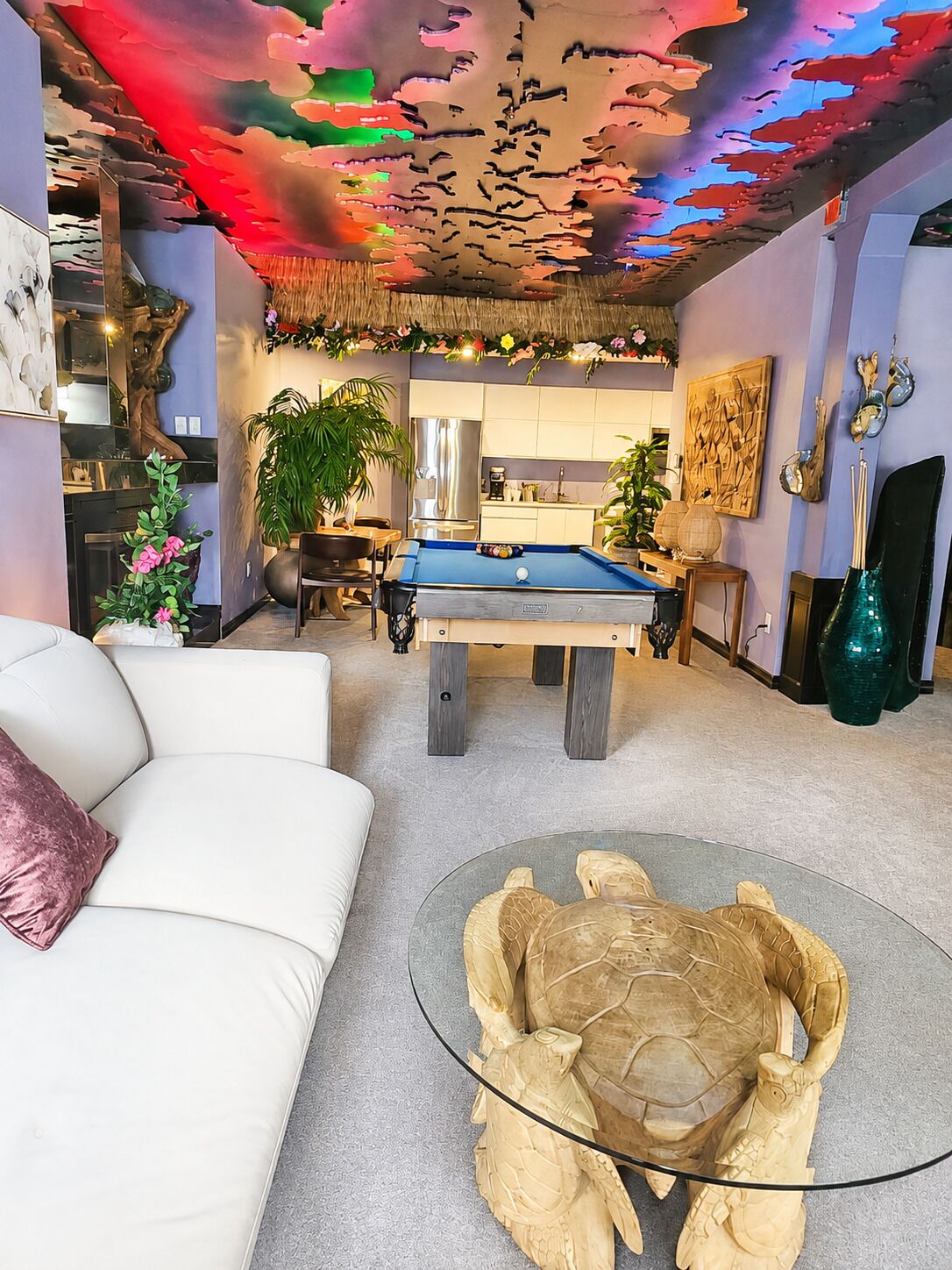 En opholdsstue med billardbord, sofa, sofabord i glas med en skildpaddeskulptur og et farverigt loftsmaleri.