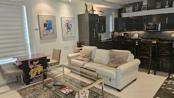A modern living room with a glass coffee table, a white sofa, and a kitchen area with dark cabinetry and a microwave.
