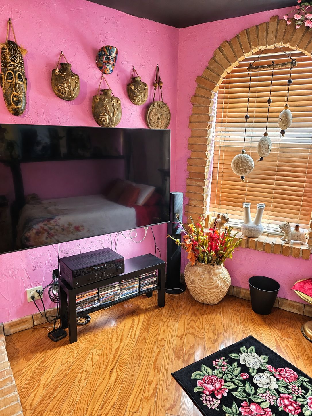 Quarto com parede rosa, cama com cabeceira preta, televisão, sistema de som e máscaras decorativas.