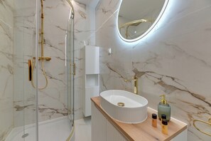 Ein modernes Badezimmer mit einer gläsernen Duschkabine, einem weißen Waschbecken mit goldfarbenen Armaturen und Marmorwänden.