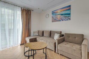 A living room with a beige sofa, a round wooden coffee table, sheer curtains, a painting of a sunset, and a clock on the wall. - Mostek 228 by Grand Apartments (Gdańsk)