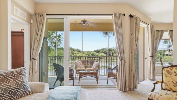 Ein Zimmer mit einer Couch, einem Sessel, einem Tisch mit einer Vase und einem Balkon mit Blick auf Grünflächen.
