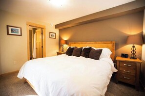 A neatly made bed with white linens and several dark pillows, a wooden headboard, a bedside table with a lamp, and a small drawer unit.