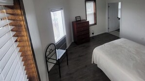 Un dormitorio con cama, cómoda, silla y escritorio con su correspondiente silla. Se observa una ventana con persianas, una puerta y una rejilla en el techo.