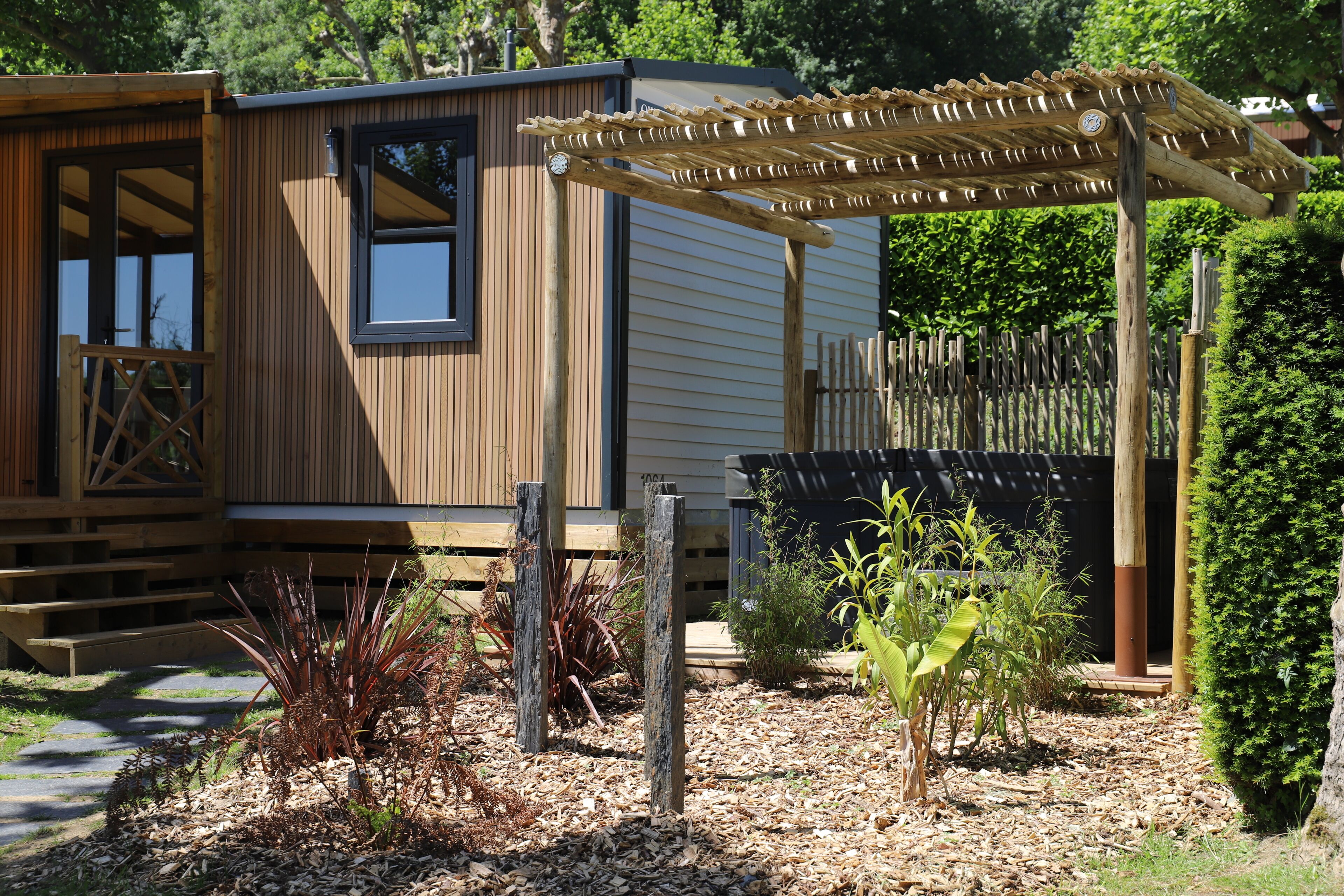 A wooden cabin with a pergola, a window, and a small garden area with plants and mulch.