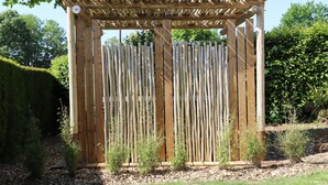 Una estructura de pérgola de madera con listones verticales, rodeada por un jardín con arbustos pequeños y un seto.