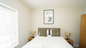A bedroom with a bed, two bedside tables, a nightstand, a wooden wardrobe, a window with blinds, and a wall-mounted abstract art piece. - Church House by Guestz (Filton)
