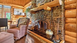 A log cabin interior with a stone fireplace, wooden walls, a cushioned chair, and a window with a view. -  Waterfront Log Cabin for Nature Lovers,moments with family friends, Spa Sleep 8 (Wayerton)