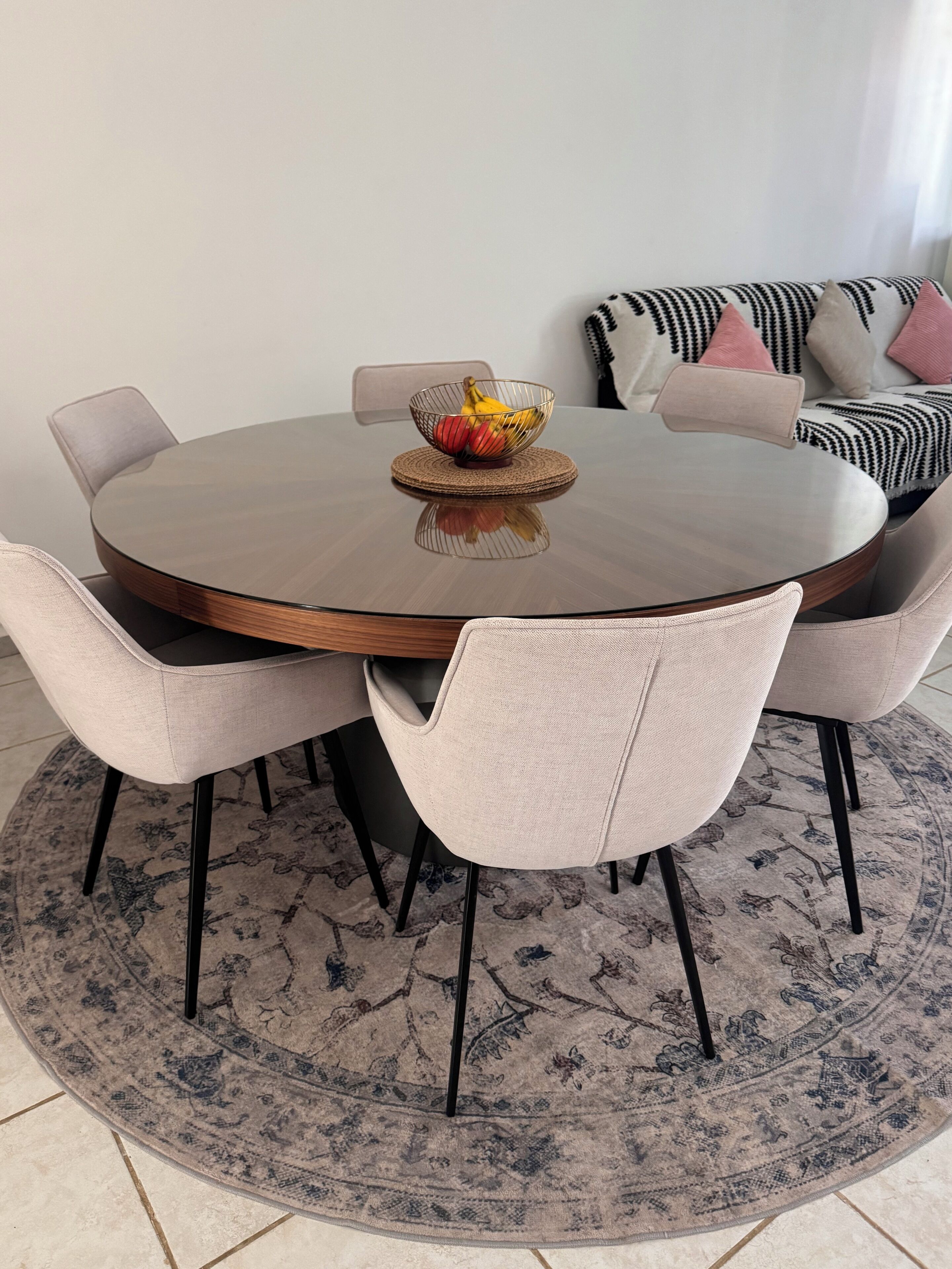 Une table de salle à manger ronde, surmontée d’un set de table en fibres tressées, entourée de six chaises rembourrées aux pieds noirs.