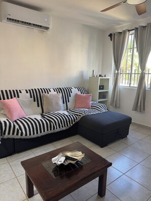 A living room with a sofa, ottoman, coffee table, air conditioning unit, ceiling fan, window with curtains, and a white cabinet. - CASA FLAMINGO - Authenticly Mexican, Peaceful 4-bedroom house in Puerto Morelos (Puerto Morelos)
